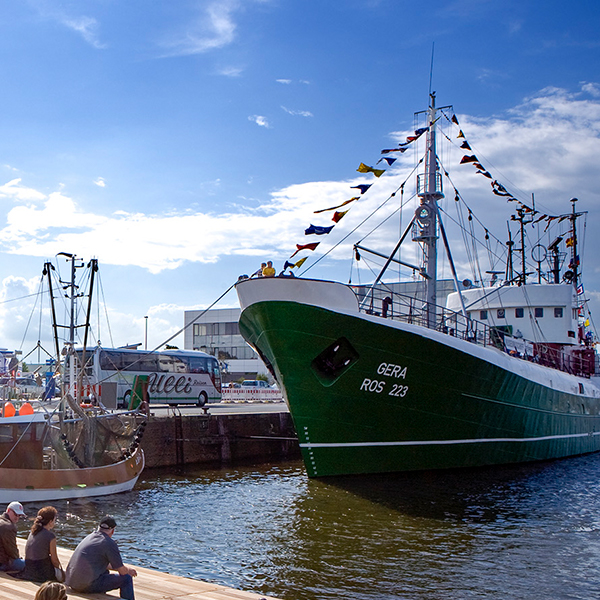 Drei Personen sitzen am Wasser vor einem Schiff.